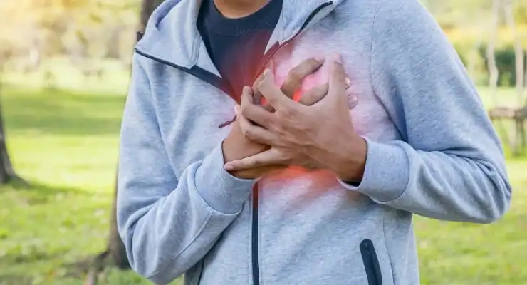 A person in a gray hoodie clutching their chest with both hands, with a red highlighted area indicating heart pain, standing in a grassy outdoor setting.
