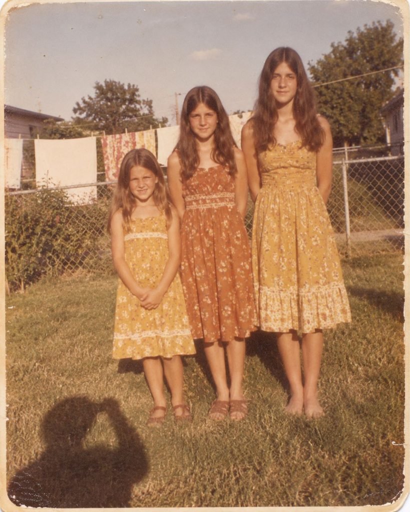 Three sisters in matching sundresses standing stiffly in a row for a single 1975 backyard snapshot